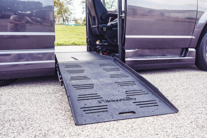 Gray minivan equipped with a ramp and other technology for a person in a wheelchair. The middle sliding doors are open with the wheelchair ramp lowered.