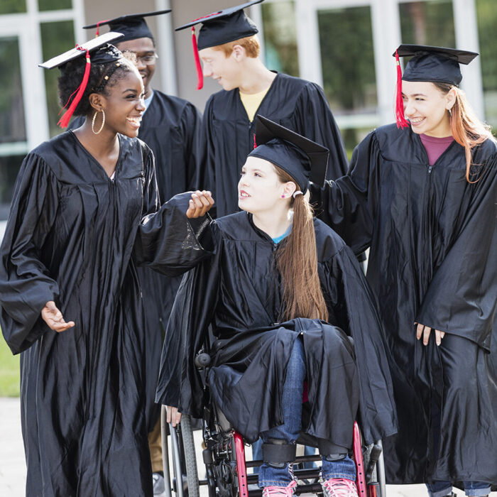 A multi-ethnic group of high school graduates, with one girl in a wheelchair, in their graduation gowns conversing together in front of the school building. 