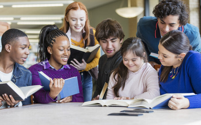 A diverse group of seven teenagers at a table in the library, reading and studying together. 