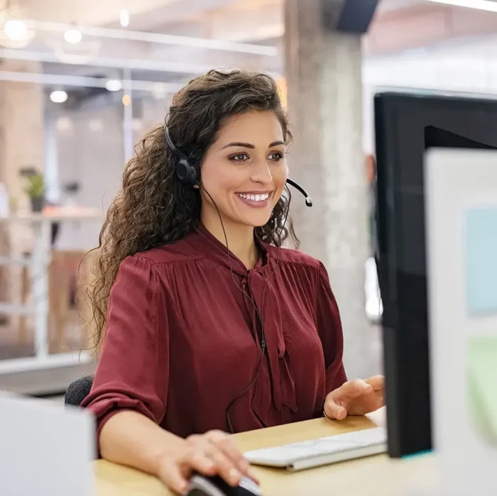Smiling woman wearing a headset is working at a computer and answering phone calls.