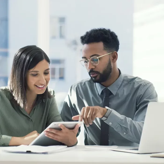 Black male with glasses wearing blue shirt and tie meets and converses with white female wearing green shirt at a desk while both look at a tablet.