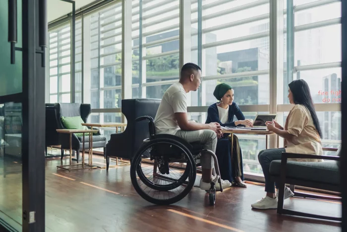 A man in a wheelchair and a woman sit at a round table in discussion with a woman.