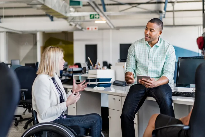 Female worker in a wheelchair sharing ideas with her male coworker, who is holding a digital tablet, in an office setting.