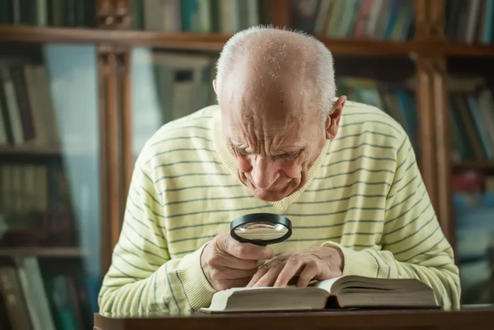 An elderly gentlemen uses a magnifying glass to read a book.