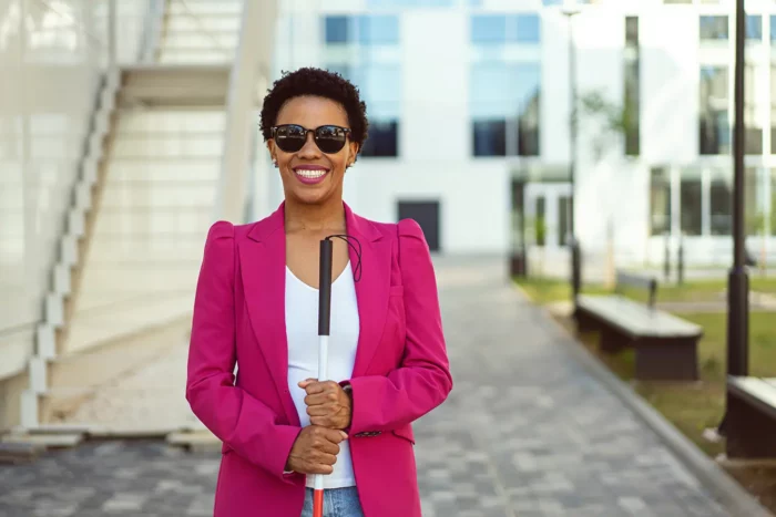 A confident mixed-race woman, with a visual impairment, stands in front of an office building while holding white cane.