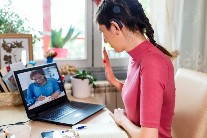 Two women meet virtually using a laptop computer. One woman hasa cochlear implant and is waving to the Black woman on her computer screen.