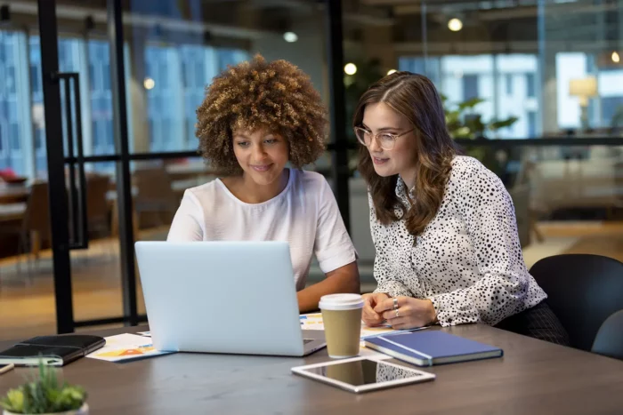 A mixed ethnic group of two women look intently at a laptop computer together. There is paperwork, coffee and technology on the table.