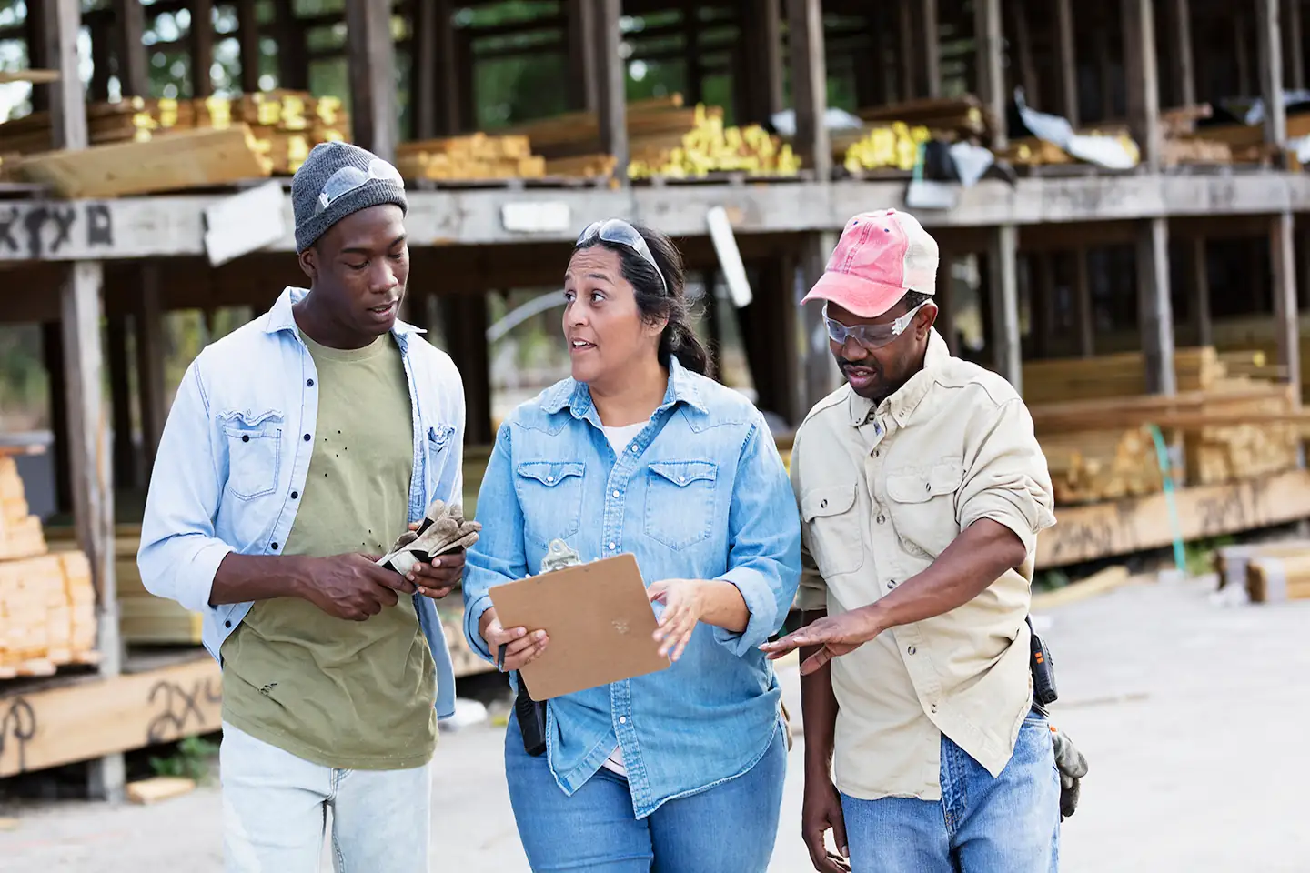 A multiracial group of three people work in a lumberyard. The manager is a Hispanic woman who is holding a clipboard. She is in the middle, looking toward a younger Black man, talking and smiling as he looks down at the clipboard.
