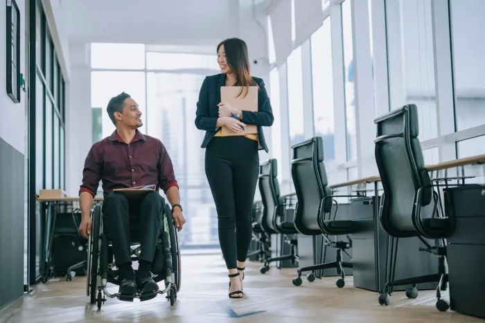 Two racially diverse individuals, one is a male in a wheelchair and the other is female, converse as they proceed down a corridor.